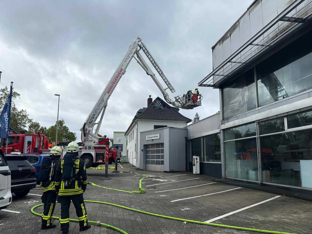 Großbrand in einer Autowerkstatt an der Kölner Straße 13 Mit dem Gelenkmast werden verbliebene Glutnester im Dachbereich aufgespürt und abgelöscht. Einsatzkräfte sichern den Bereich während der Arbeiten ab.