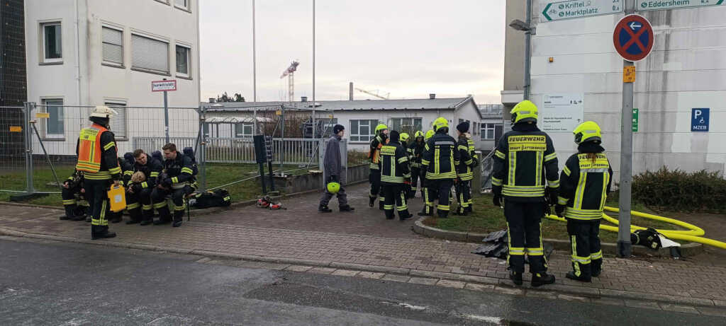 Einsatzkräfte der Feuerwehr bei den Nachlöscharbeiten an dem betroffenen Gewerbeobjekt. Der Einsatzbereich ist teilweise mit Löschschaum abgedeckt, Fahrzeuge und Geräte stehen rund um das Gebäude bereit.