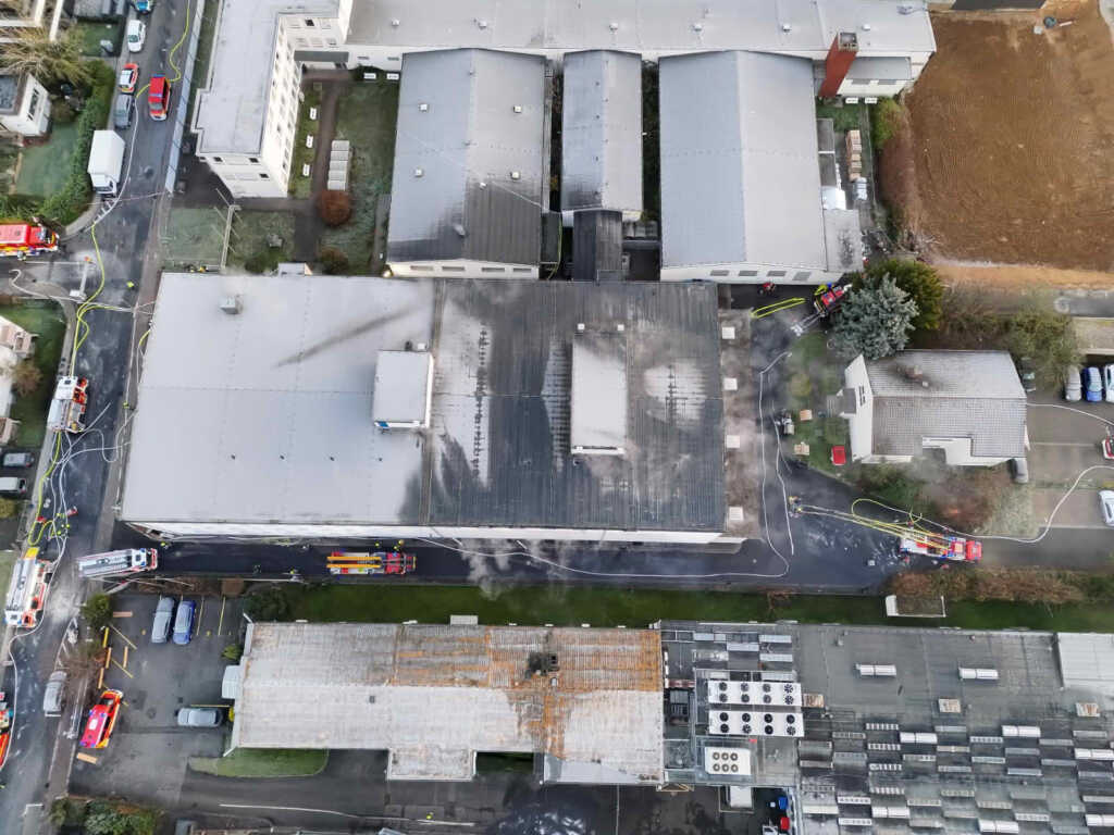 Einsatzkräfte der Feuerwehr bei den Nachlöscharbeiten an dem betroffenen Gewerbeobjekt. Der Einsatzbereich ist teilweise mit Löschschaum abgedeckt, Fahrzeuge und Geräte stehen rund um das Gebäude bereit.